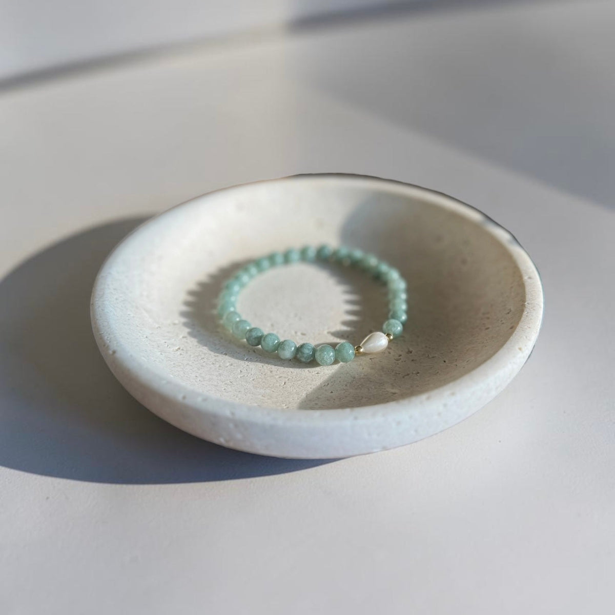 A beaded stretch bracelet in light mint green and white, displayed inside a small, round, textured ceramic dish on a neutral light gray surface.