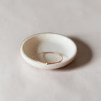 A delicate brown wire bangle bracelet with a tiny gold bead accent, displayed inside a small, round, off-white ceramic dish on a neutral beige surface.