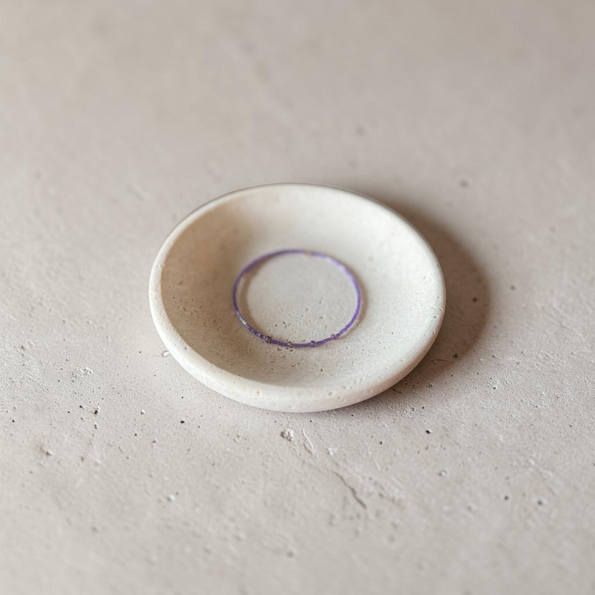 A small round ceramic dish with a textured matte finish, containing a thin purple wire bracelet, set against a neutral concrete background.