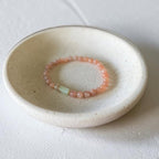 A delicate beaded bracelet in shades of coral pink and light green rests inside a small, round, textured off-white ceramic dish on a matching light surface.