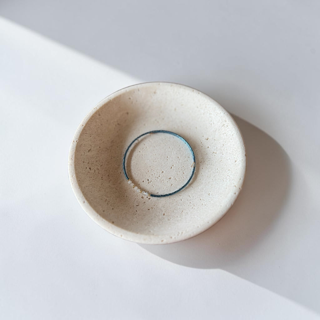 A minimalist ceramic dish with a speckled cream glaze, featuring a thin wire bracelet its center, photographed from a top-down angle on a white surface with soft shadows.