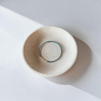 A minimalist ceramic dish with a speckled cream glaze, featuring a thin wire bracelet its center, photographed from a top-down angle on a white surface with soft shadows.
