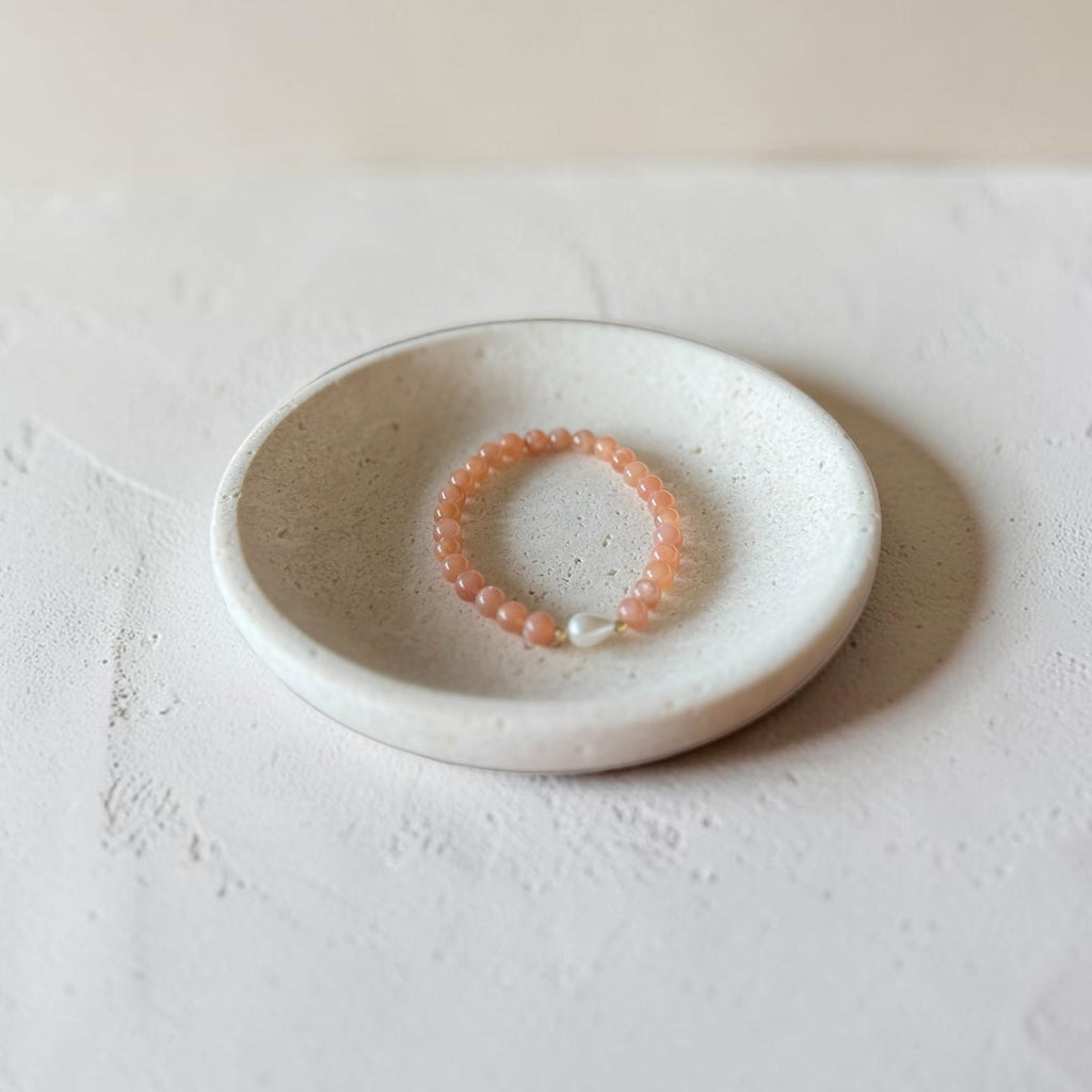 A peach-orange beaded bracelet with a white pearl accent, displayed inside a small round white ceramic dish on a textured white surface.