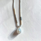 A beaded necklace with a teardrop-shaped iridescent shell pendant, laid flat on a textured off-white surface. The necklace features white beads, small gold-toned spacer beads, and black-and-white striped beads.