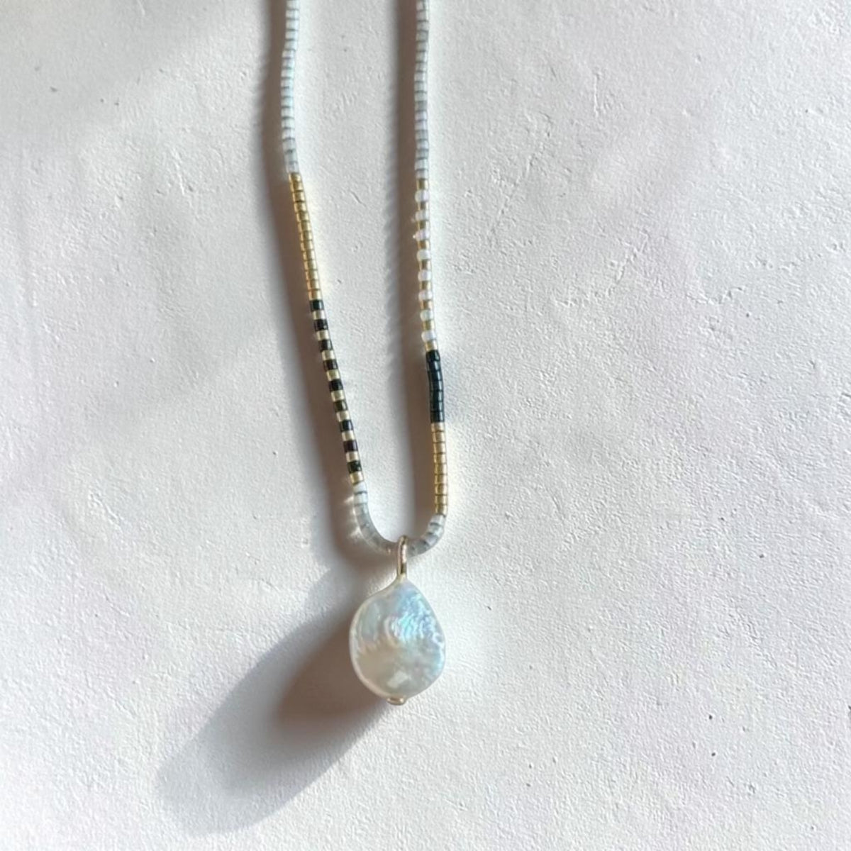A beaded necklace with a teardrop-shaped iridescent shell pendant, laid flat on a textured off-white surface. The necklace features white beads, small gold-toned spacer beads, and black-and-white striped beads.