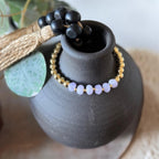 A beaded bracelet with pale lavender-purple oval beads and small gold spacer beads, draped around a matte black ceramic vase. The photo is styled with a tassel and green leaf props on a soft neutral background.