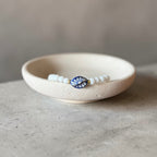 A beaded bracelet with white round beads and a central blue and white floral enamel charm, displayed inside a small textured cream ceramic dish on a gray concrete surface.