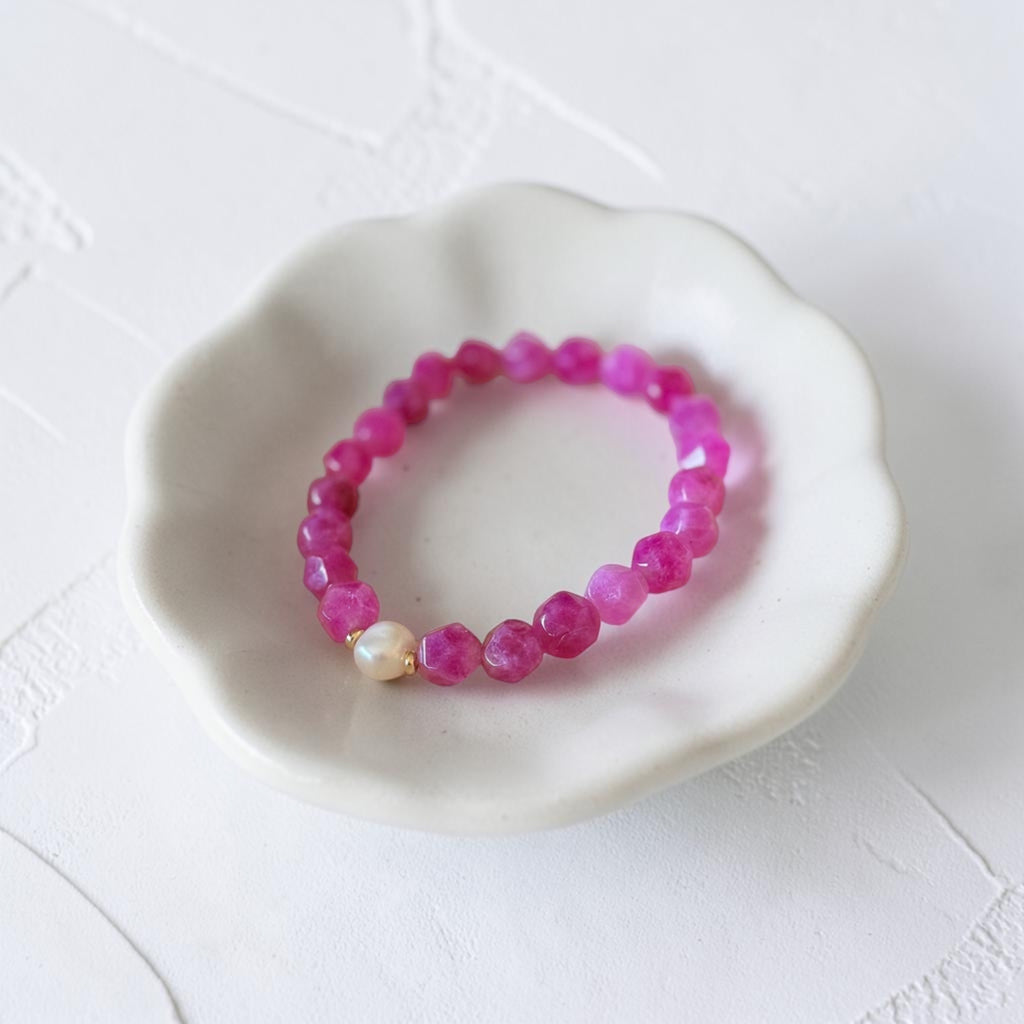 A pink beaded stretch bracelet with a small white pearl accent, displayed on a white ceramic flower-shaped dish against a textured white background.