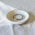A beaded stretch bracelet with small translucent brown beads and a white stone accent, displayed inside a small round ceramic dish on a white fabric surface.