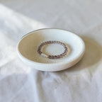 A beaded stretch bracelet with small translucent brown beads and a white stone accent, displayed inside a small round ceramic dish on a white fabric surface.