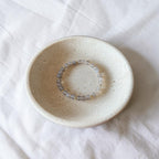 A beaded stretch bracelet with translucent gray-blue beads and tiny gold spacer beads, displayed inside a small speckled ceramic dish on a white fabric background.