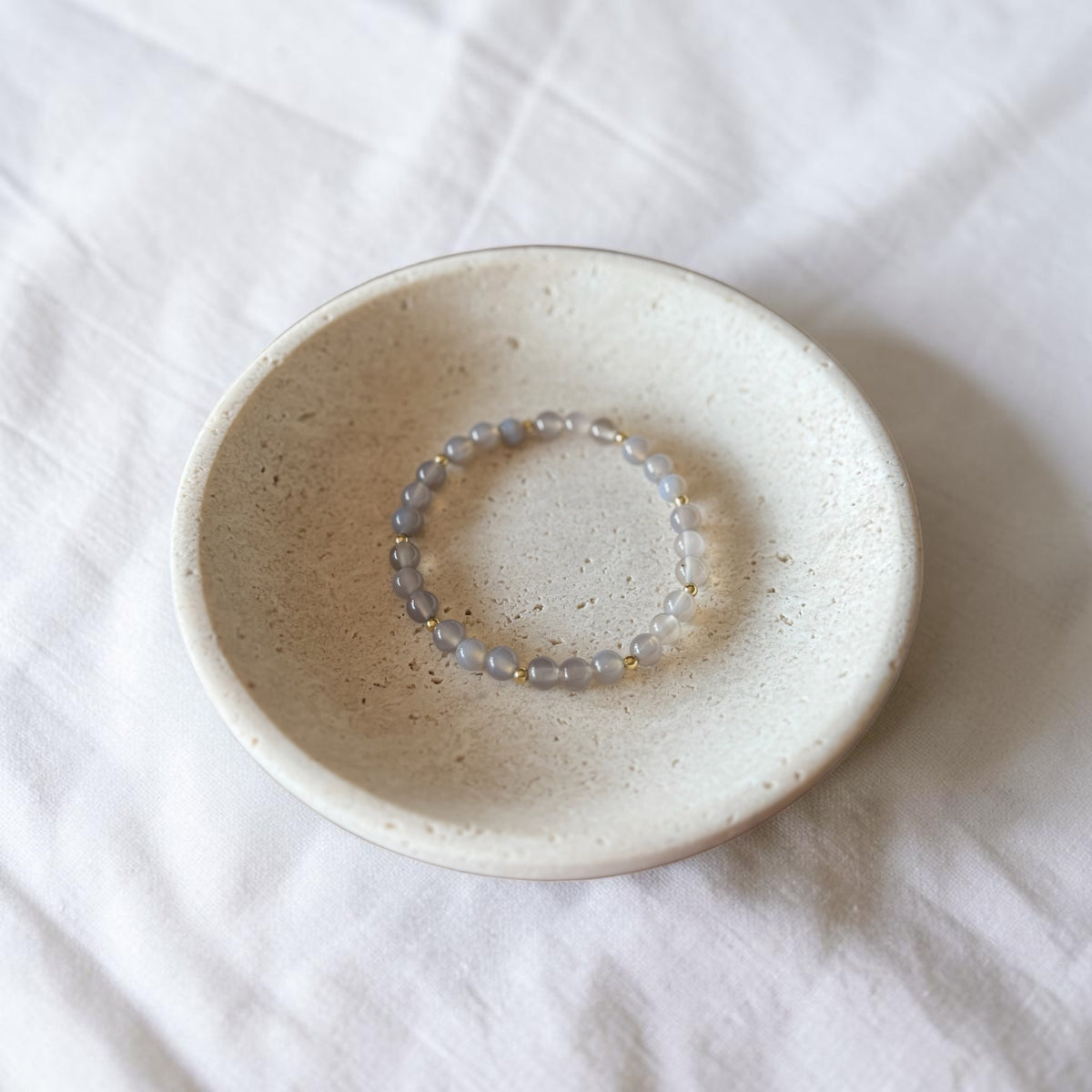 A beaded stretch bracelet with translucent gray-blue beads and tiny gold spacer beads, displayed inside a small speckled ceramic dish on a white fabric background.