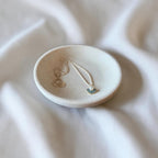 A small round ceramic jewelry tray holding a delicate necklace with a small blue and white fan-shaped charm, set against a softly draped white fabric background.