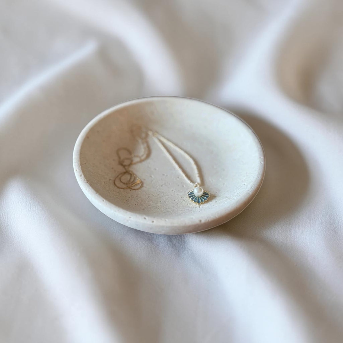 A small round ceramic jewelry tray holding a delicate necklace with a small blue and white fan-shaped charm, set against a softly draped white fabric background.