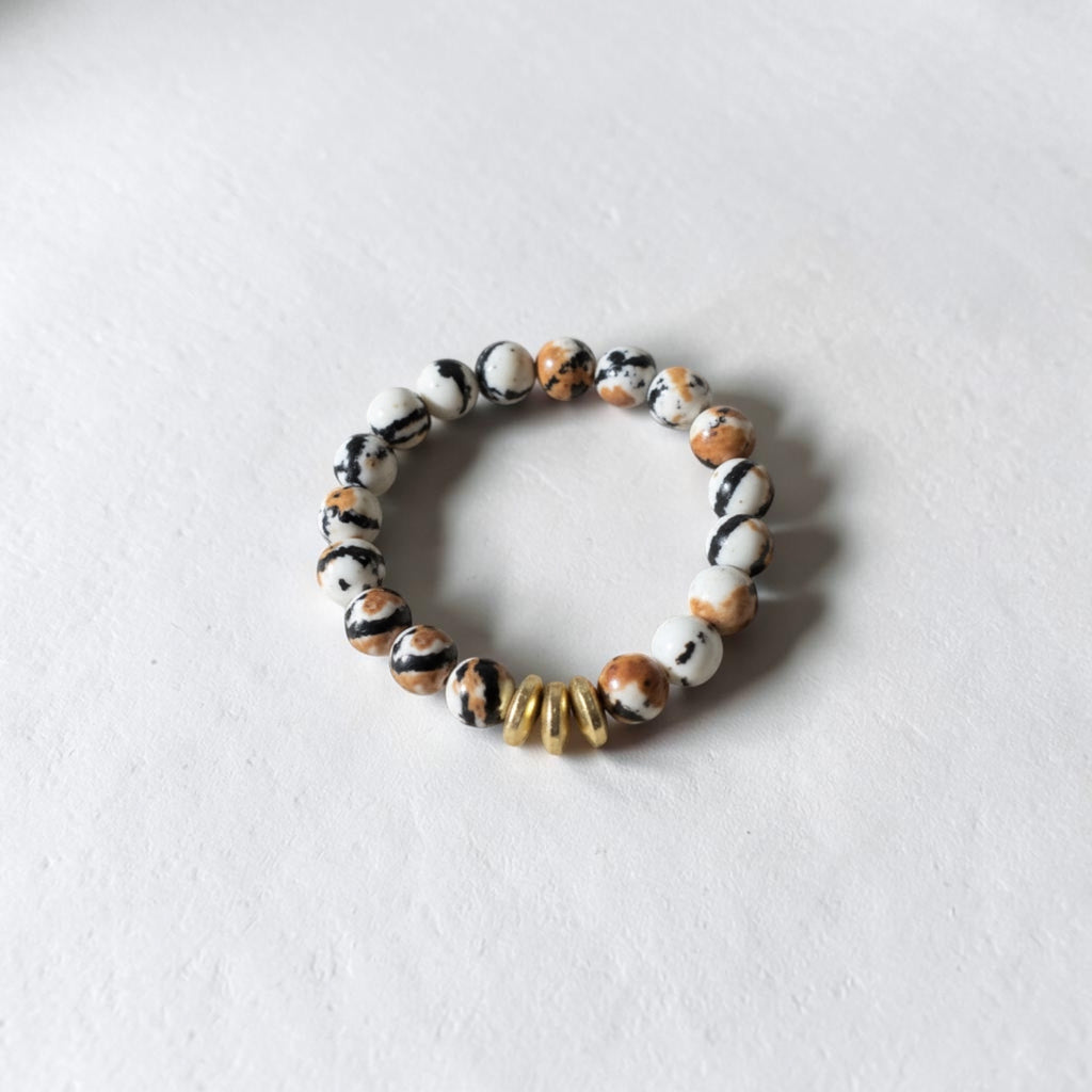 A beaded stretch bracelet featuring marbled white, black, and brown beads, accented by three small gold spacer beads, shown on a light textured background.