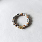 A beaded stretch bracelet featuring marbled white, black, and brown beads, accented by three small gold spacer beads, shown on a light textured background.
