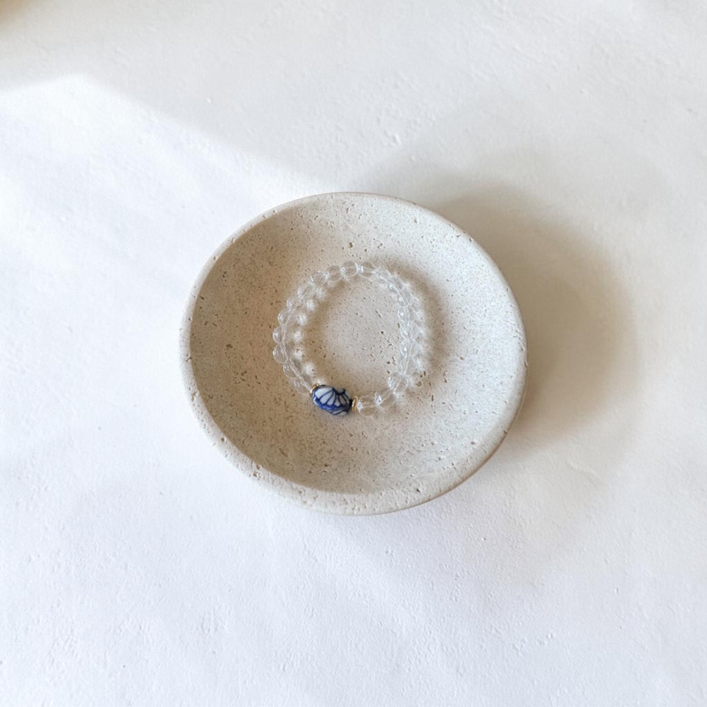 A beaded bracelet with a small blue and white patterned bead, displayed inside a small speckled ceramic dish on a white surface.