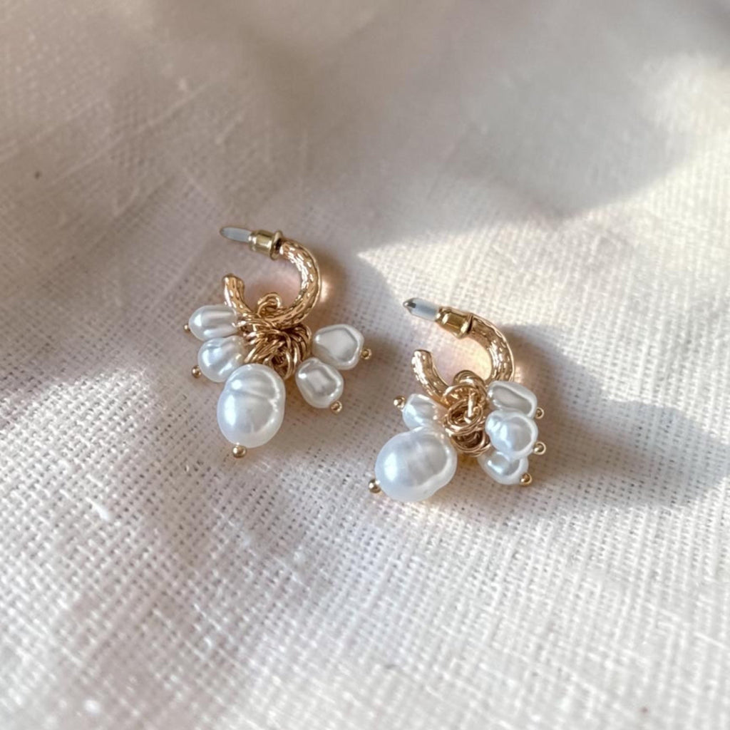 A pair of gold hoop earrings with dangling white baroque pearls, displayed on a textured cream linen fabric under soft natural light.
