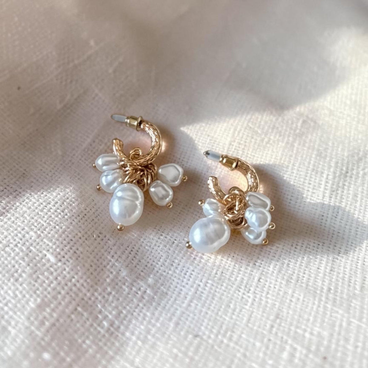 A pair of gold hoop earrings with dangling white baroque pearls, displayed on a textured cream linen fabric under soft natural light.