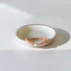 A peach-beige beaded bracelet with a small gold charm, displayed on a small, round, speckled ceramic dish against a bright white background with soft shadows.
