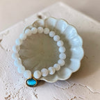 A beaded bracelet with white round beads and a small oval light blue charm in a gold-tone setting, displayed on a scalloped off-white ceramic dish over a textured beige surface.