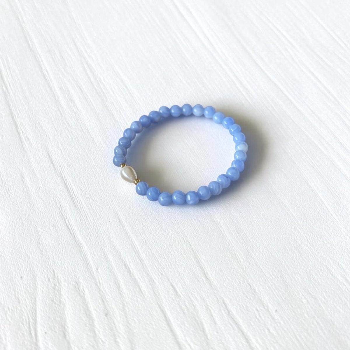 A light blue beaded stretch bracelet with a small white bead accent, displayed on a white textured background.