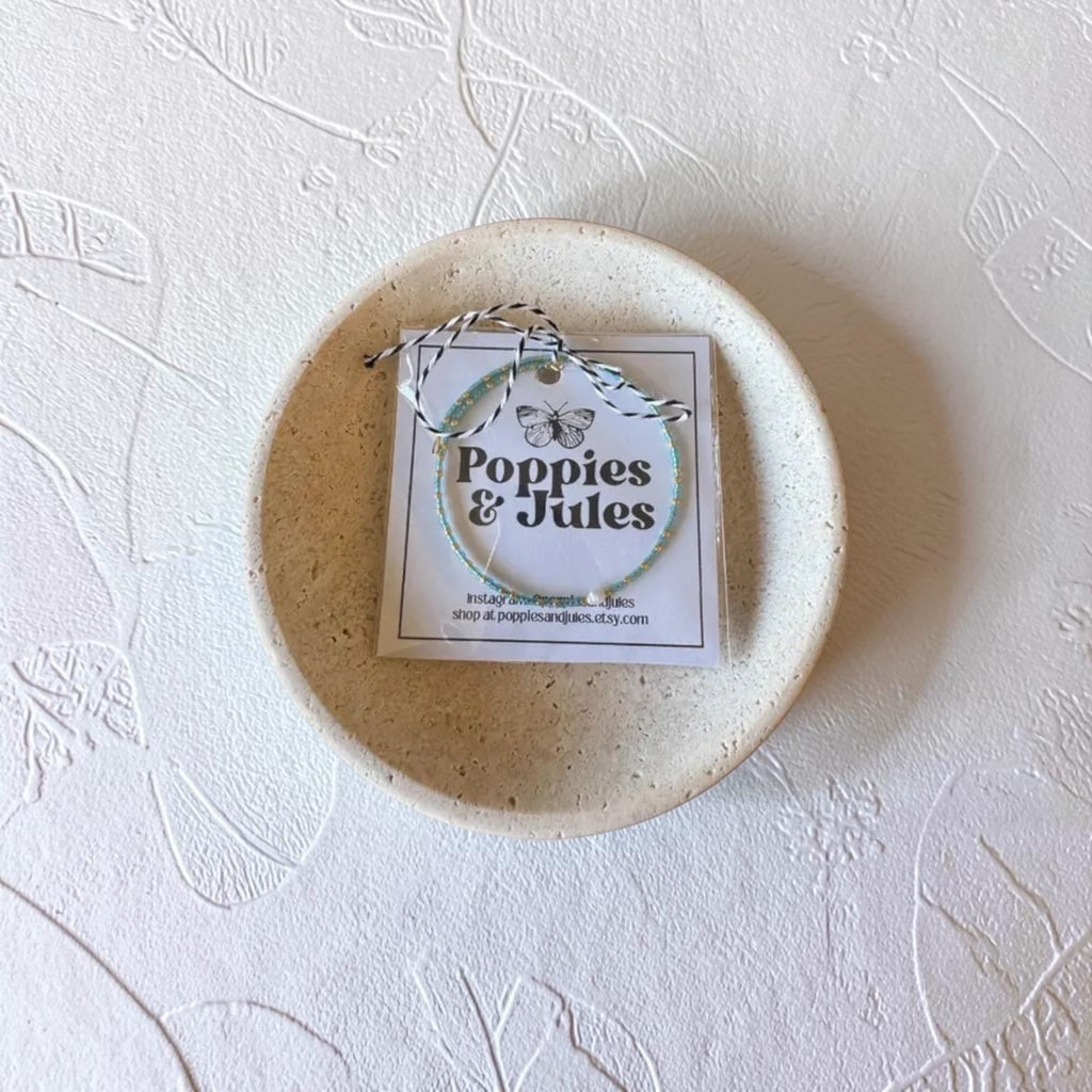 A small round ceramic dish in a speckled cream-beige glaze sits on a textured white surface. Inside the dish is a square white card with the brand name 'Poppies & Jules' printed in black, tied with a string.