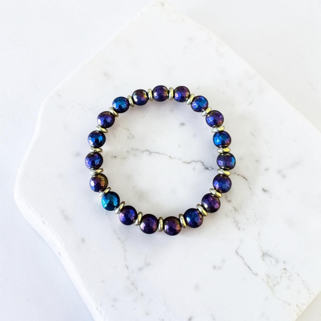A beaded stretch bracelet with iridescent dark blue and purple round beads separated by small gold spacer rings, displayed on a white marble slab.