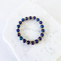 A beaded stretch bracelet with iridescent dark blue and purple round beads separated by small gold spacer rings, displayed on a white marble slab.