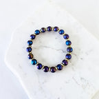 A beaded stretch bracelet with iridescent dark blue and purple round beads separated by small gold spacer rings, displayed on a white marble slab.