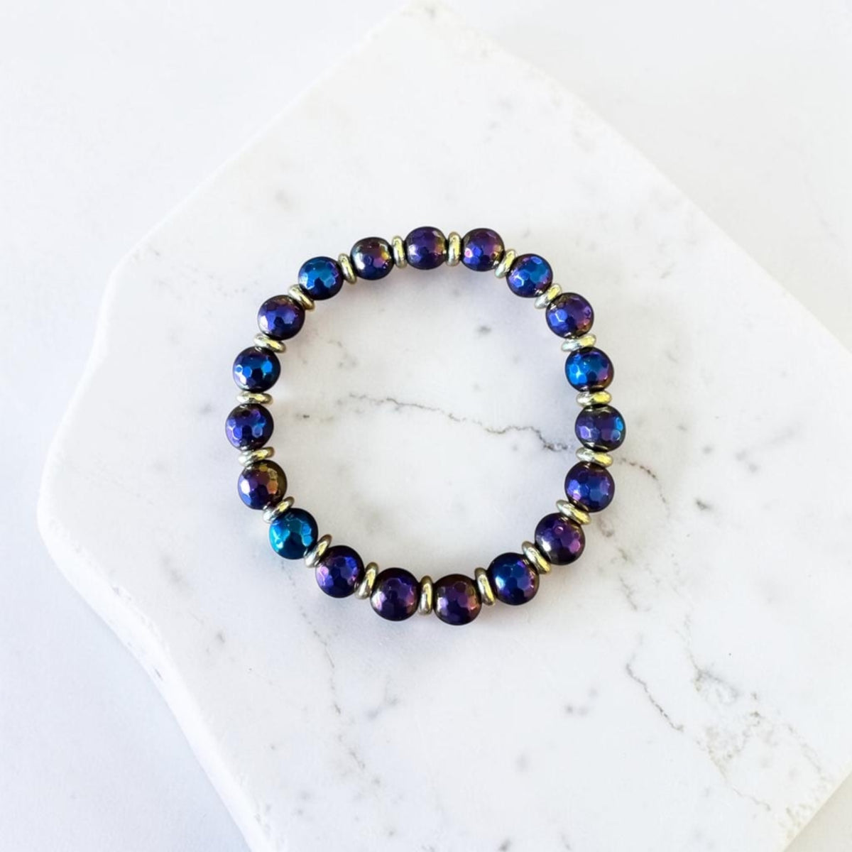 A beaded stretch bracelet with iridescent dark blue and purple round beads separated by small gold spacer rings, displayed on a white marble slab.