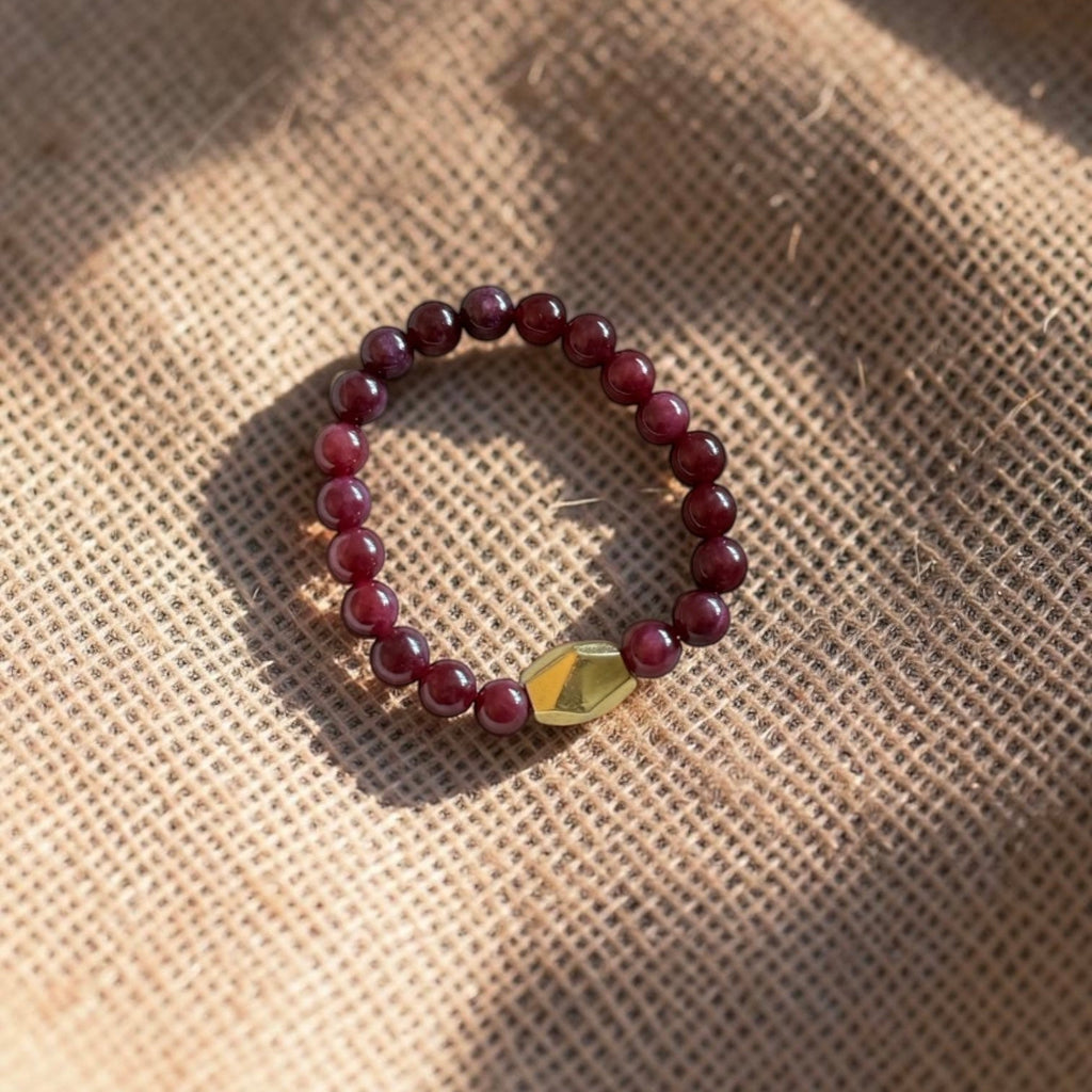 A beaded stretch bracelet with dark red marbled beads and a faceted gold accent bead, resting on a textured beige burlap surface with soft shadows.