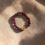 A beaded stretch bracelet with dark red marbled beads and a faceted gold accent bead, resting on a textured beige burlap surface with soft shadows.