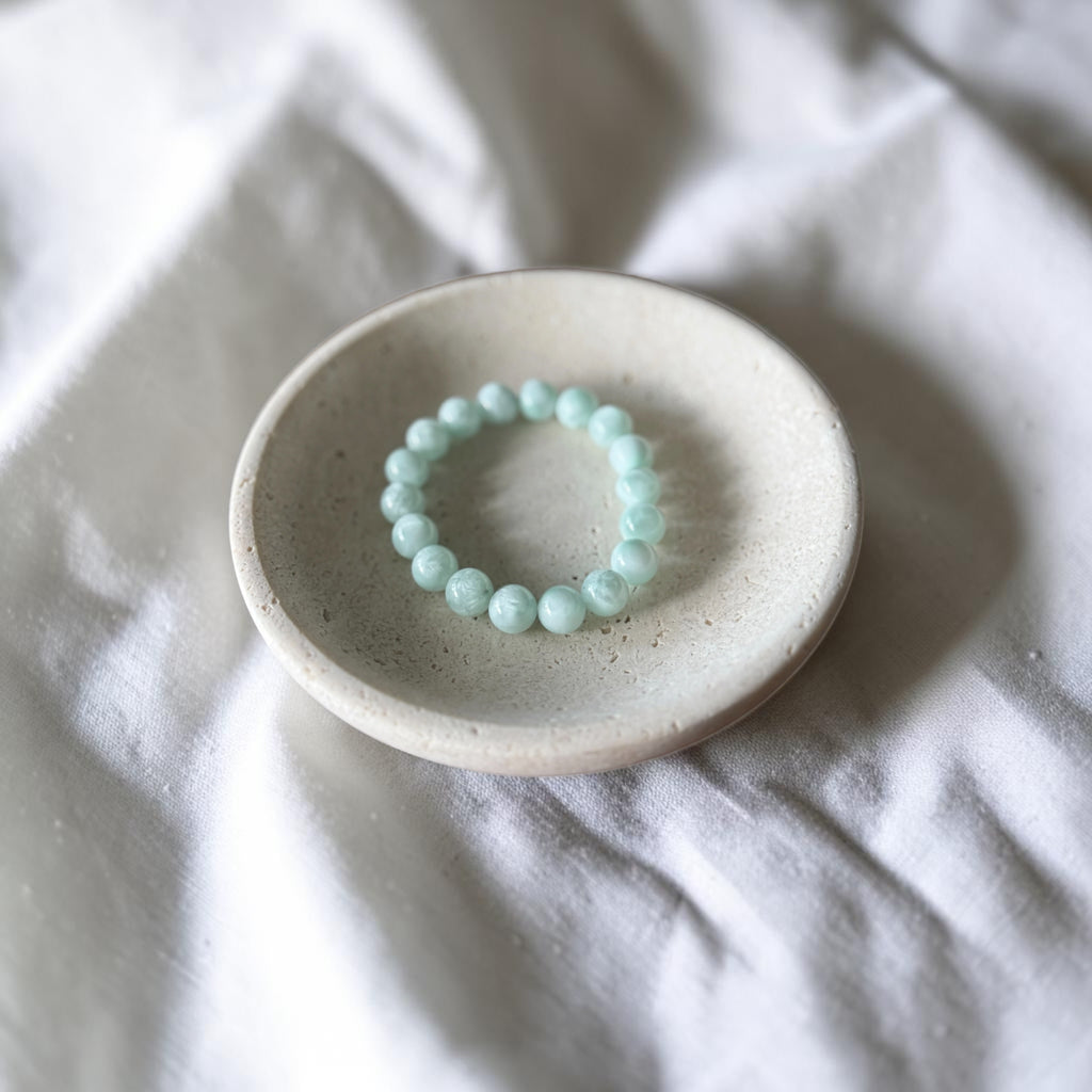 A light mint green beaded bracelet displayed on a small, round, speckled ceramic dish over a soft, white fabric background.