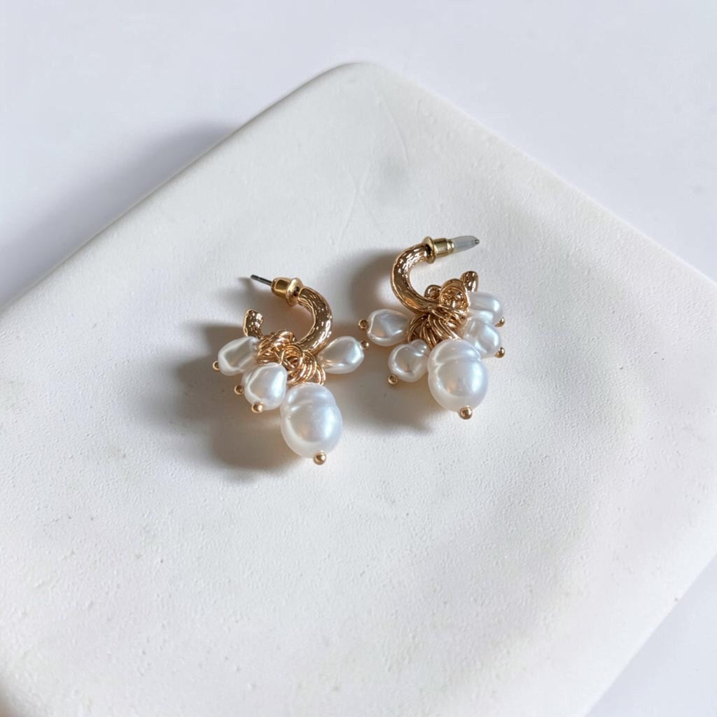 A pair of gold hoop earrings with white pearl clusters, displayed on a white textured square tile.