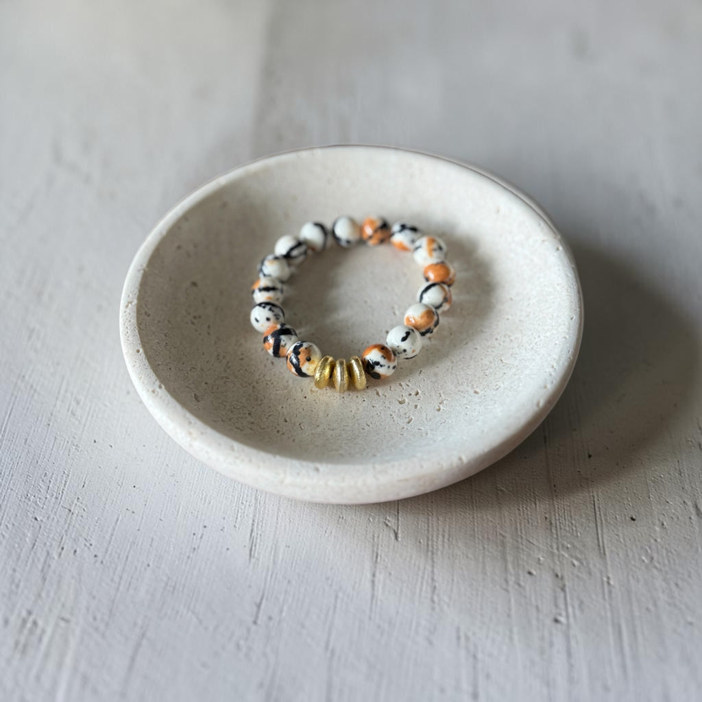 A beaded stretch bracelet with white, black, and orange marbled beads and a small gold accent bead, displayed on a small round ceramic dish over a light gray textured surface.