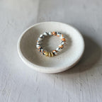 A beaded stretch bracelet with white, black, and orange marbled beads and a small gold accent bead, displayed on a small round ceramic dish over a light gray textured surface.
