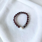 A beaded stretch bracelet featuring faceted dark burgundy beads and a single white pearl accent, displayed on a soft white fabric background.