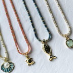 Four delicate beaded necklaces with gold-tone pendants are arranged diagonally on a textured white surface. The necklaces feature coral pink, metallic blue, and cream beads, with pendants including a gold fish, a gold turtle, a blue and white seashell with a pearl, and a light green stone.