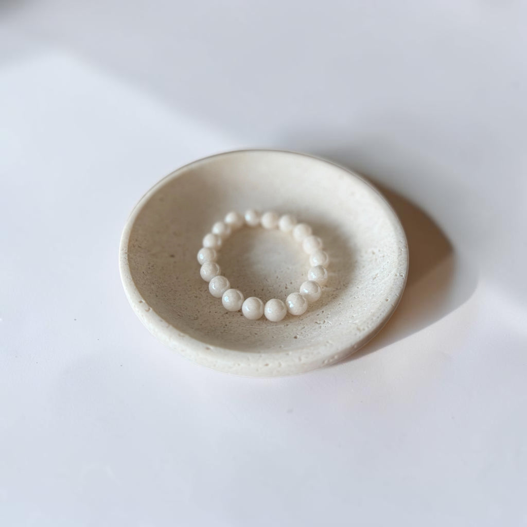 A beaded pearl bracelet displayed inside a small, round, textured ceramic dish on a clean white surface with soft shadows.