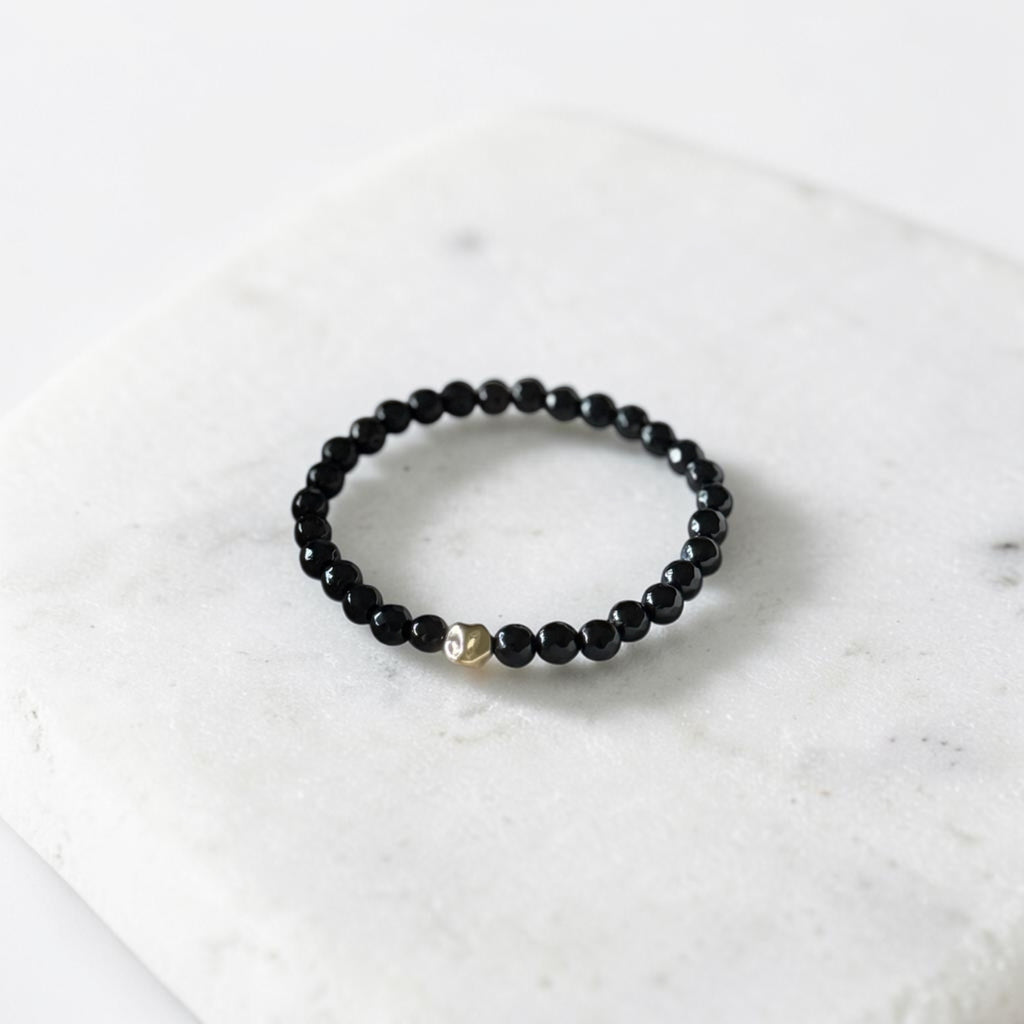 A beaded stretch bracelet with small black faceted beads and a single gold-toned accent bead, displayed on a white marble slab.