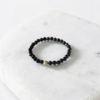 A beaded stretch bracelet with small black faceted beads and a single gold-toned accent bead, displayed on a white marble slab.