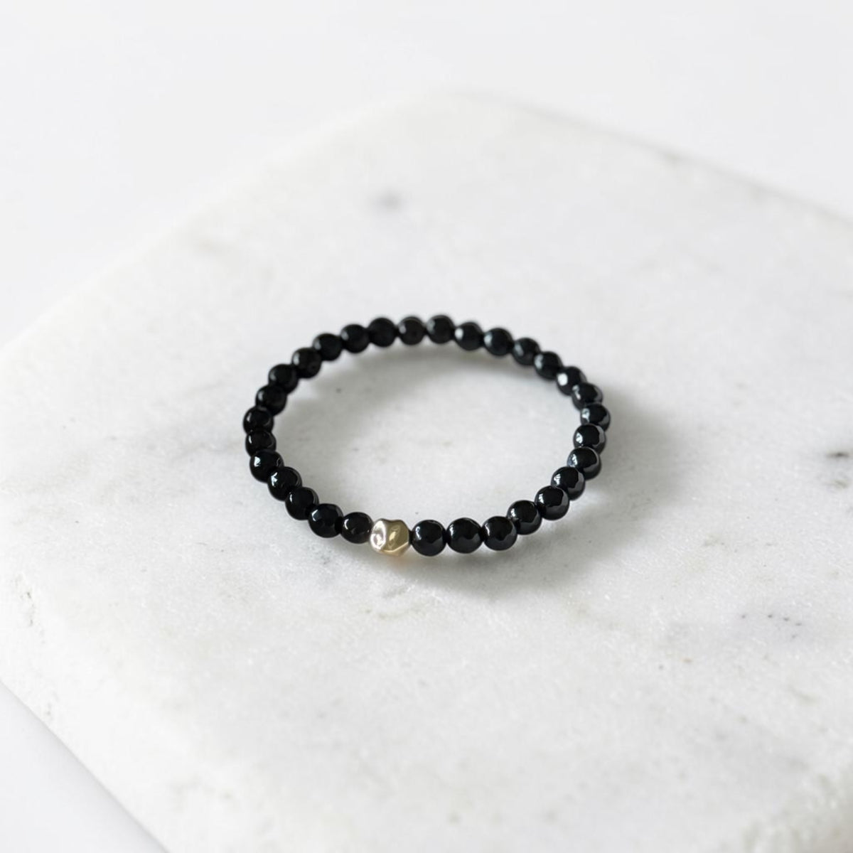 A beaded stretch bracelet with small black faceted beads and a single gold-toned accent bead, displayed on a white marble slab.