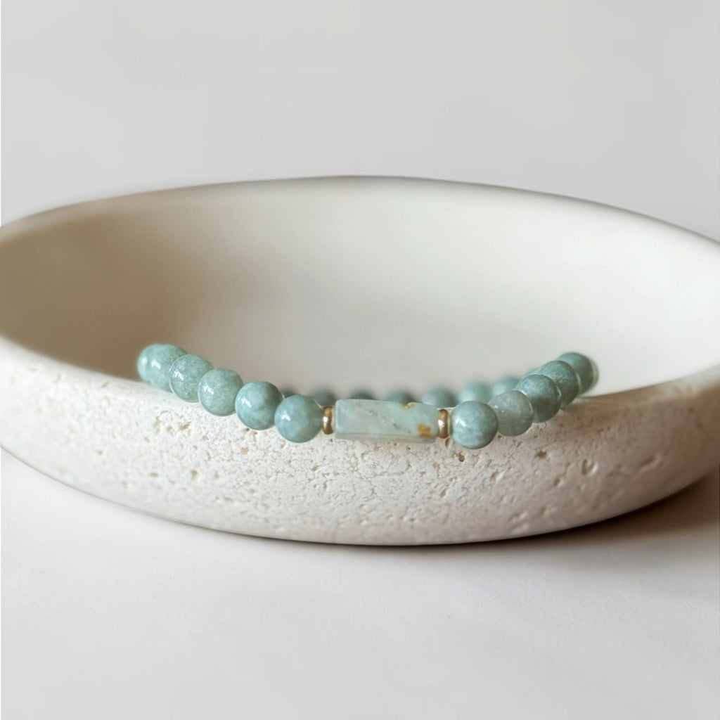 A beaded stretch bracelet with light blue-green round beads and a central rectangular stone, displayed on a textured off-white ceramic dish against a neutral light gray background.