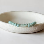 A beaded stretch bracelet with light blue-green round beads and a central rectangular stone, displayed on a textured off-white ceramic dish against a neutral light gray background.