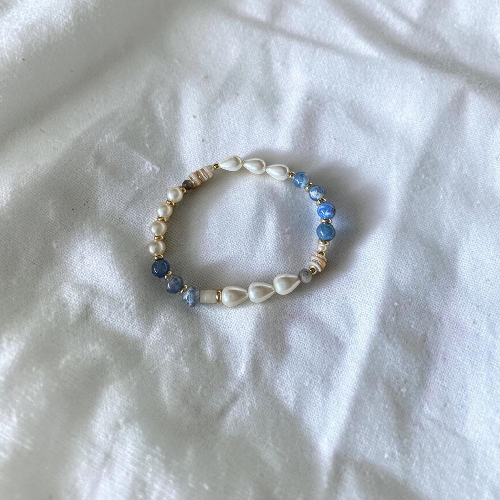 A beaded stretch bracelet featuring white cowrie shells, blue faceted beads, and small metallic spacer beads, displayed on a wrinkled white fabric background.