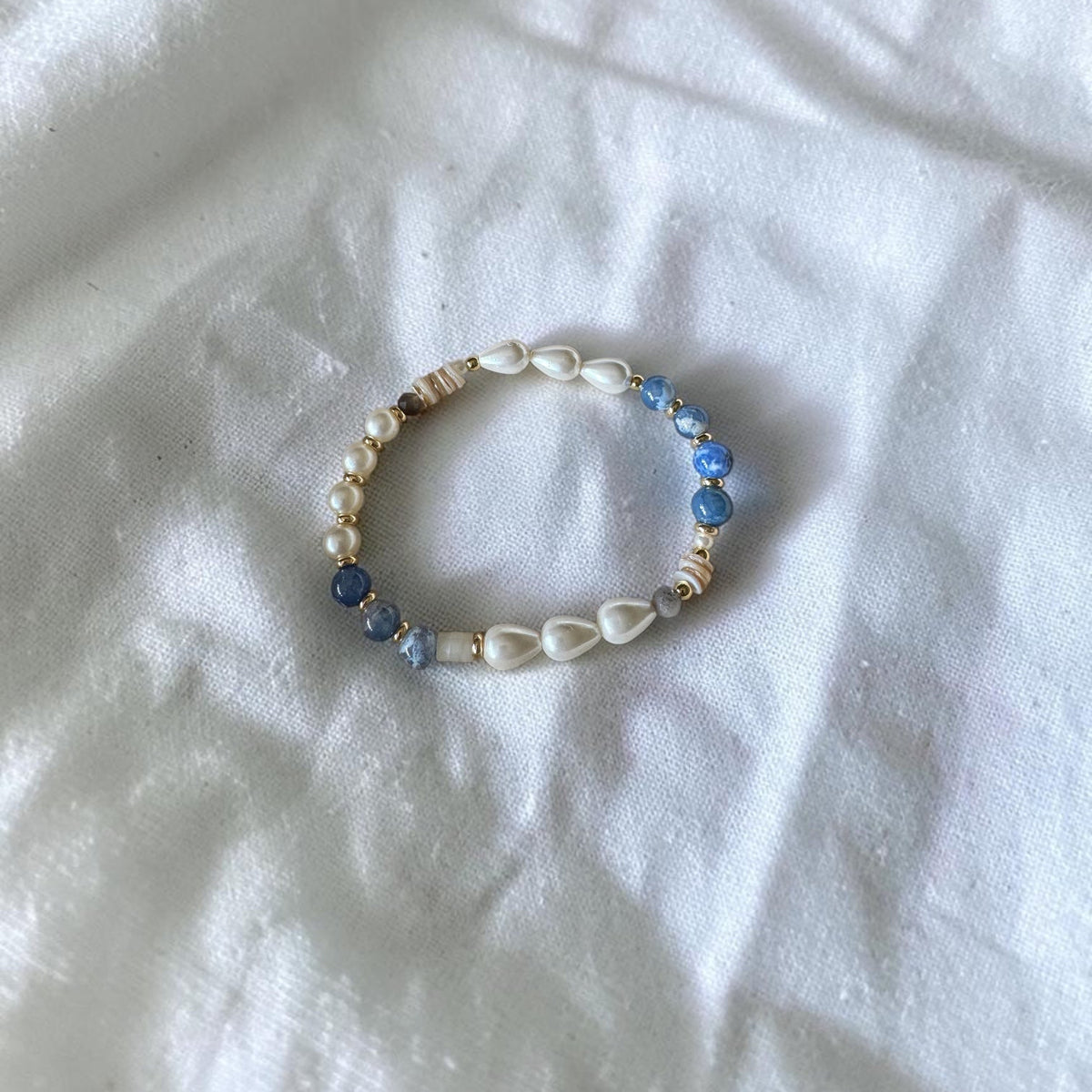 A beaded stretch bracelet featuring white cowrie shells, blue faceted beads, and small metallic spacer beads, displayed on a wrinkled white fabric background.