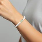 A close-up of a person's wrist wearing a beaded bracelet with white round beads and a single pale green rectangular accent bead, against a soft light gray background.