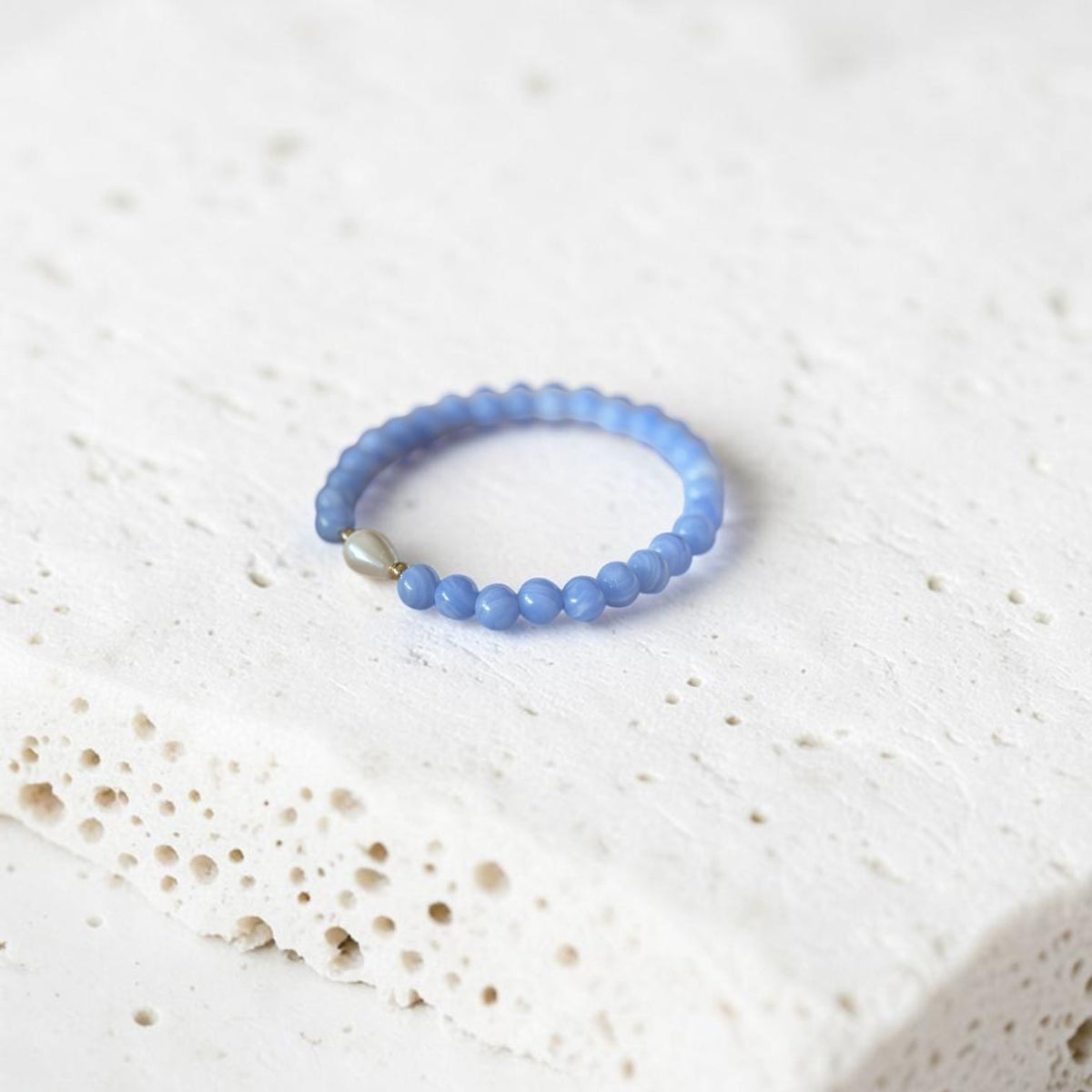A light blue beaded bracelet with a small silver-toned accent bead, resting on a textured white stone slab.