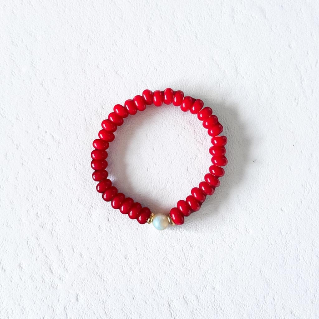 A red beaded bracelet with a small white pearl accent, displayed on a textured white background.